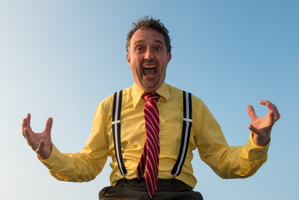 Marcel Harmsen mid-performance in a yellow shirt and red striped tie, against open sky