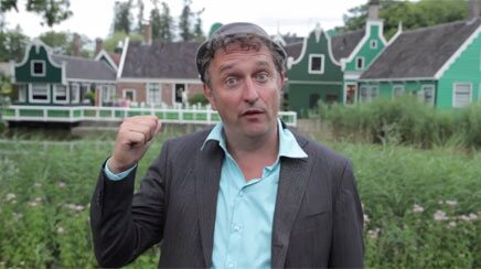 Marcel Harmsen filmed outdoors in front of green Dutch houses, mid-line