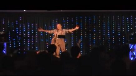 Marcel Harmsen alone on a lit stage in front of a company audience, arms thrown wide