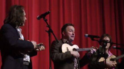 Marcel Harmsen singing at a microphone with two musicians against a red curtain