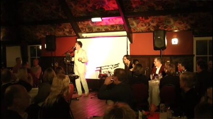Marcel Harmsen performing beside a projection screen to seated guests at round tables