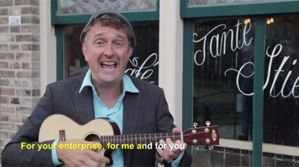 Marcel Harmsen singing to camera with a ukulele, a subtitle across the foot of the frame