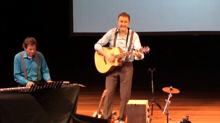 Marcel Harmsen playing guitar on a theatre stage with a keyboard player behind him